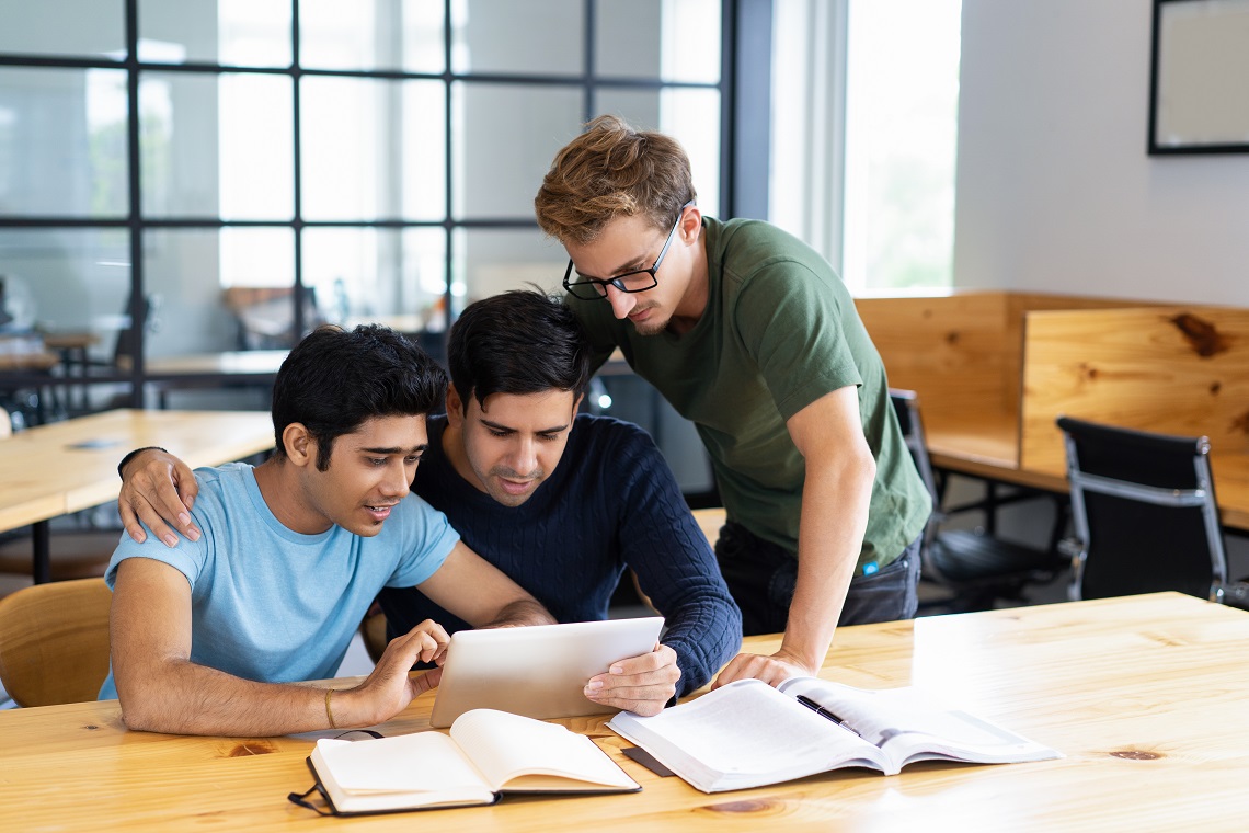 Focused students browsing on tablet computer and talking. Young men studying, standing and sitting at desk in classroom or library. Education and friendship concept.